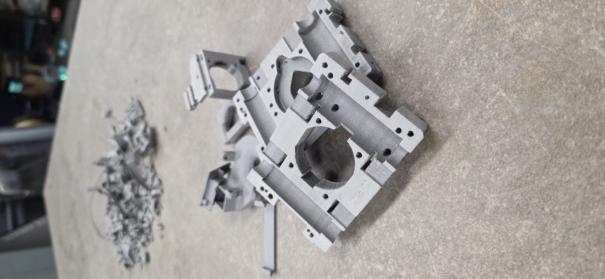 Grijs geprint frameonderdeel met meerdere bevestigingsgaten en een grote ronde uitsparing, net van de printer met resten steunmateriaal ernaast.